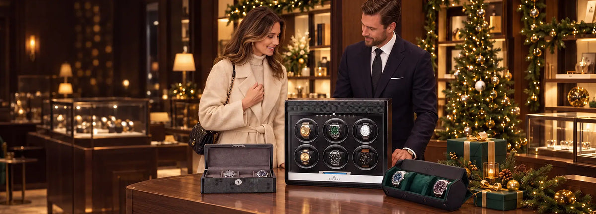 A man and a woman stand in a luxury watch winder boutique next to a large black 6-watch automatic watch winder display case. The man wears a dark navy suit, and the woman wears a beige trench coat.