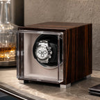 Single watch winder in glossy ebony Macassar wood with a clear display case holding a silver automatic chronograph watch.